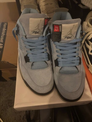 Jordan 4 university blue  - Jordan 4 university blue  med ovandel i mocka och meshdetaljer. Skorna har högt skaft, ljusblå skosnören och svarta samt grå detaljer på sidorna och sulan. Tungan har en grå patch med röd och svart detalj. Perfekta för dig som gillar streetwear.