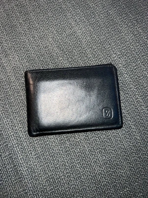 Svart skinnplånbok med enkel design - Stilren svart plånbok i skinn med minimalistisk look och diskret logga i hörnet. Perfekt storlek för kort och sedlar, smidig att ha i fickan. Passar dig som gillar klassiska accessoarer med en modern touch.