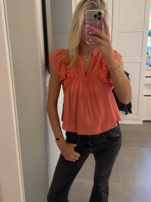 Korallfärgad blus med volang - Supersöt korallfärgad blus med volangdetaljer och v-ringning. Blusen är luftig och har korta ärmar. Perfekt till jeans eller kjol 