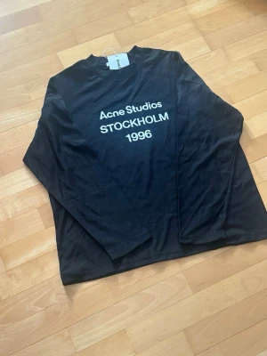 T-shirt manches longues Acne Studios 1996  - Helt ny långärmad t-shirt från Acne Studios i svart, med det ikoniska vita trycket ”Acne Studios STOCKHOLM 1996” på framsidan. Tröjan är tillverkad i mjuk bomull och har snygga slitna detaljer vid kragen och fållen, vilket ger den en unik, ingången känsla.   Skick: 10/10  Storlek: M  Färg: Svart Snabb och omsorgsfull frakt
