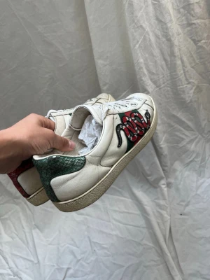 Vita sneakers med orm och färgdetaljer - Unika vita sneakers med broderad orm i rött, svart och grönt på sidan. Hälpartiet har kontrasterande detaljer i röd och grön mockaimitation med reptilmönster. Klassisk låg modell med vita snören och sula i gummi. Perfekta för dig som vill sticka ut.