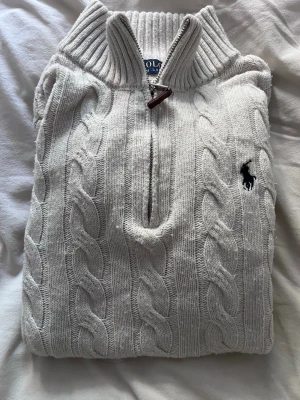 Vit kabelstickad tröja Polo Ralph Lauren (170) - Vit kabelstickad tröja med halv dragkedja och hög krage från Polo Ralph Lauren. Tröjan har ett broderat mörkblått logotypmärke på bröstet och ribbstickade detaljer vid krage och ärmslut. Perfekt för en klassisk och stilren look.