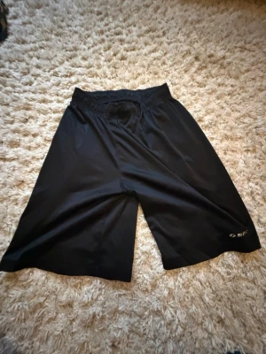 Svarta träningsshorts från Select - Svarta träningsshorts från Select med elastisk midja och luftig passform. Tillverkade i ett lätt och snabbtorkande syntetmaterial, perfekta för sport och aktivitet. Diskret logga på ena benet.