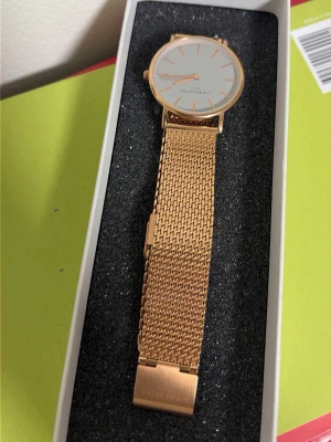 Guldig klocka från Liebeskind Berlin - Stilren klocka från Liebeskind Berlin med mesh-armband i guld och rund vit urtavla. Minimalistisk design med tunna visare och index. Perfekt accessoar för dig som gillar en clean look och vill ha något som sticker ut lite extra.