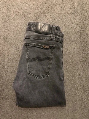 Gråa skinny jeans från Nudie Jeans - Säljer ett par gråa skinny jeans från Nudie Jeans med klassisk femficksmodell och svarta sömmar på bakfickorna. Jeansen har en svart patch i midjan och är tillverkade i mjuk denim med stretch. Perfekta för en avslappnad och trendig look. Storleken är W30/L32. Pris kan diskuteras. Skriv vid minsta fråga. 