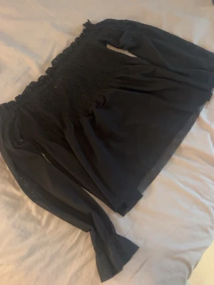 Svart offshoulder topp Gina Tricot - Trendig svart offshoulder topp från Gina Tricot med långa ärmar och smockad överdel. Toppen har volangdetalj upptill och är tillverkad i ett lätt, tunt material som ger en luftig känsla. Perfekt för dig som vill ha en stilren och chic look .