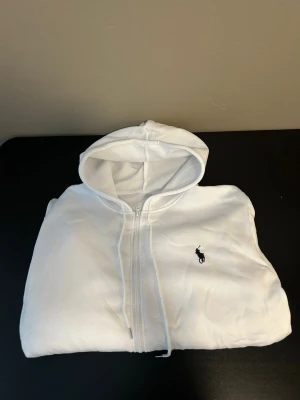 Vit hoodie med dragkedja från Polo Ralph Lauren - Snygg vit hoodie med dragkedja från Polo Ralph Lauren. Klassisk design med huva och det ikoniska mörkblå logotrycket på bröstet. Tillverkad i mjukt material som känns skönt mot huden. Perfekt för en clean och stilren look.