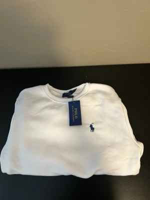 Vit sweatshirt från Polo Ralph Lauren - Klassisk vit sweatshirt från Polo Ralph Lauren med rund halsringning och den ikoniska blå logotypen broderad på bröstet. Tröjan har en avslappnad passform och är tillverkad i mjukt material som passar perfekt till en casual stil.