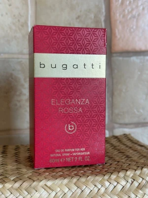 Bugatti röd Eau de Parfum 60 ml - Bugatti Eau de Parfum i en elegant röd glasflaska med svart lock och silverdetalj. Flaskan rymmer 60 ml och har en stilren, modern design med märkets logga på framsidan. Perfekt för dig som gillar exklusiva dofter och snygg förpackning. Oöppnad! Tog ut flaskan enbart för att fota.