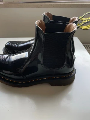 Svarta lackade Dr.Martens - Säljer ett par svarta Dr.Martens i glansigt lackmaterial med klassisk resår på sidorna och grov sula. Skorna har gul söm runt sulan och dragflik bak. 
