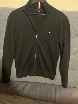 Svart ziptröja från Tommy Hilfiger - Snygg svart ziptröja från Tommy Hilfiger med liten logga på bröstet och ribbad hög krage med röd, vit och blå rand. Tröjan har långa ärmar och ribbade muddar vid ärmslut och nederkant. Perfekt till lager på lager och enkel att matcha.