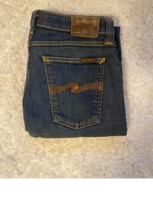 Mörkblå jeans från Nudie Jeans - Snygga mörkblå jeans från Nudie Jeans med klassisk femficksdesign och kontrastsömmar på bakfickorna. Jeansen har en smal passform och är tillverkade i denim med läderpatch i midjan. Perfekta för en trendig och avslappnad stil. Storlek 30 men sitter mer som en 28-29