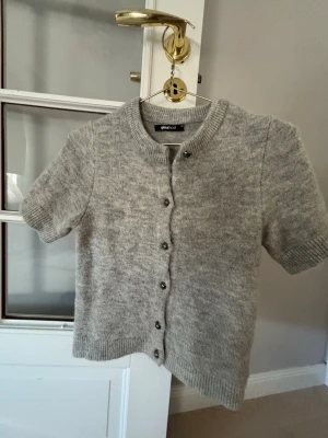 Grå kortärmad kofta med knappar - Mysig grå kofta med korta ärmar och rund halsringning. Koftan har knäppning framtill med små knappar, tröjan e från Gina trikot och är i fint skick.