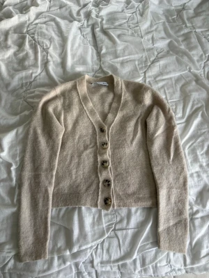Beige kofta med knappar - Mysig beige kofta med v-ringning och fem stora knappar framtill. Koftan har långa ärmar och ribbade muddar vid ärmslut och nederkant. Tillverkad i ett mjukt, stickat material som passar perfekt för lager på lager.