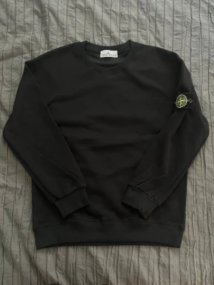 Stone island tröja. - Säljer en helt ny svart långarmad Medium tröja från Stone Island med den klassiska loggan på vänster ärm. Tröjan har rund halsringning, ribbade muddar vid ärmslut och nederkant samt en clean och stilren look. Perfekt för dig som gillar streetwear och vill ha något enkelt men ändå ikoniskt.