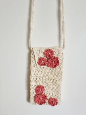 Virkad mobilväska med blommor - Handgjord virkad axelremsväska i beige med rosa virkade blommor som dekoration. Väskan har ett långt axelband, stängs lätt med tryckknapp. Perfekt för mobilen eller småsaker när du vill ha händerna fria. Material 100% bomull och stärkt för att hålla formen.