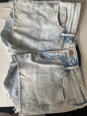 Ljusblå jeansshorts från Hollister - Snygga ljusblå jeansshorts från Hollister med slitna kanter och klassisk femficksdesign. Shortsen har en avslappnad passform och är perfekta för varma dagar. De har en knappstängning och cool tvättad look.