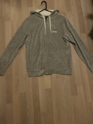 Grå hoodie med dragkedja från BOSS - Mysig grå hoodie från BOSS med dragkedja framtill och vita snören i huvan. Tröjan har fickor på sidorna och en liten BOSS-logga på bröstet. Perfekt för chill dagar och enkel att matcha med jeans eller mjukisbyxor.