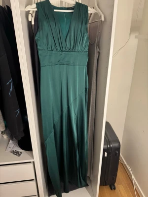Mörkgrön långklänning med slits - Elegant mörkgrön långklänning i ett glansigt material med v-ringad hals och bred midjedetalj. Klänningen är ärmlös och har en snygg slits framtill som ger en extra touch. Perfekt för dig som vill sticka ut på nästa tillställning!