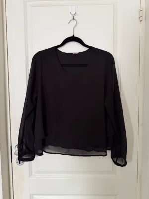 Svart transparent blus med lång ärm - Snygg svart blus i transparent chiffong med rund halsringning och långa, flödande ärmar. Blusen har ett dubbelt lager tyg framtill för en stilren look och ett lätt, luftigt fall. Perfekt att styla med jeans eller kjol för en trendig outfit.
