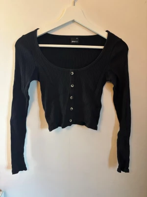 Svart ribbad croppad topp med knappar - Snygg svart croppad topp med långa ärmar och ribbad struktur. Toppen har en djup rundringning och silverfärgade knappar framtill. Perfekt att styla med höga jeans eller kjol för en trendig look.