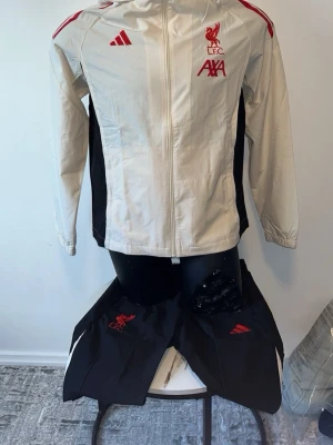 Liverpool träningsset från Adidas - Snyggt träningsset från Adidas med Liverpool FC-logga. Jackan är vit med röda detaljer, huva och dragkedja framtill. Byxorna är svarta med röda ränder och elastisk midja. Perfekt för fotbollsträning eller sportig stil.