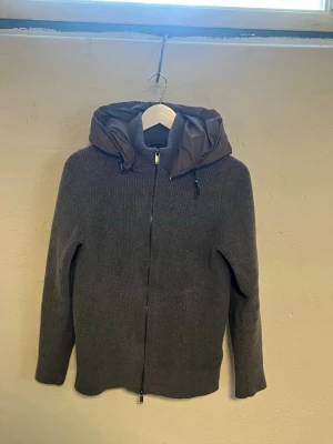 Massimo dutti cardigan  - Mysig grå stickad cardiganjacka med dragkedja framtill och en vadderad brun huva. Jackan har lång ärm och ribbad struktur, perfekt för kyliga dagar. Enkel och stilren design som passar till det mesta.