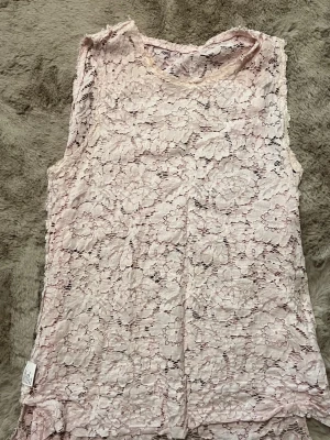 Ljusrosa ärmlös spetsblus - Säljer en ljusrosa ärmlös blus i spets med blommigt mönster. Toppen har rund halsringning och är transparent, perfekt att styla med ett linne under. Superfin till både jeans och kjol!