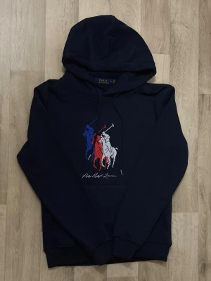 Mörkblå hoodie Polo Ralph Lauren - Snygg mörkblå hoodie från Polo Ralph Lauren med tryck av tre polospelare i blått, rött och vitt på bröstet. Klassisk huva med dragsko och magficka framtill. Perfekt för dig som gillar streetwear och vill ha en stilren look. För endast 699kr
