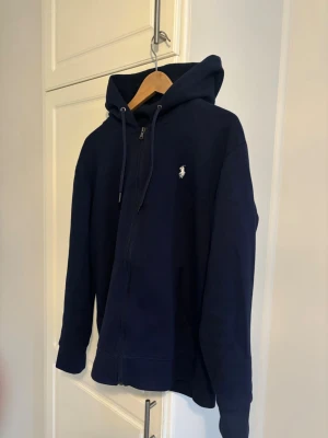 Polo Ralph Lauren  zip hoodie - Mörkblå Polo Ralph Lauren zip hoodie med dragkedja och huva, justerbara snören och fickor framtill. Klassisk passform och diskret logga broderad på bröstet. Perfekt för en chill och stilren look. Materialet ser ut att vara mjuk bomullsblandning. Ända felet är att en del av snöret har ramlat av som syns i sista bilden.