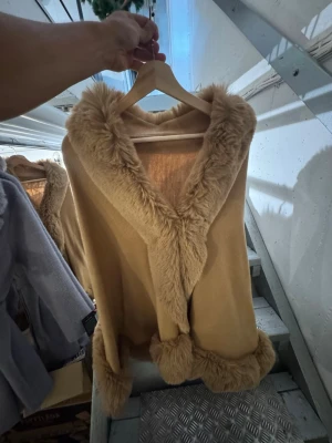 Beige pälsjacka/kofta med fuskpälsdetaljer Garoff - Snygg beige pälsväst med mjuka fuskpälsdetaljer längs krage och nederkant. Västen har en öppen design utan knäppning och ger en lyxig känsla till din outfit. Perfekt för dig som vill ha något extra till din look. Finns att hämta i Gröndal.
