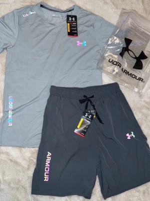 2 Under Armour träningsset t-shirt & shorts - Snygga träningsset från Under Armour med t-shirt och shorts. Finns i både ljusgrått och svart. Setet har färgglada loggor och textdetaljer, samt elastisk midja med dragsko på shortsen. Perfekt för gymmet eller löprundan. S/M båda träningsetten för 1000 kr