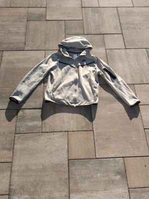 Ljusgrå nike tech XL Barnstorlek - Snygg ljusgrå hoodie från Nike i storlek barn XL. Hoodien har dragkedja framtill, huva och en ficka på ärmen. Tillverkad i mjukt material som känns skönt mot huden. Perfekt för dig som gillar sportig och avslappnad stil.