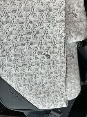 Vit handväska  - Snygg vit handväska  med det klassiska mönstret i grått och vitt. Väskan har detaljer i svart läder och präglad logotyp på hörnet. Perfekt accessoar för dig som gillar exklusiv och tidlös design.