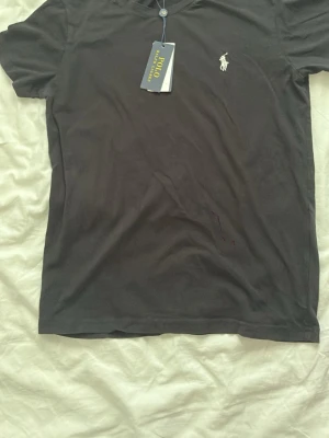 Svart t-shirt från Polo Ralph Lauren - Snygg svart t-shirt från Polo Ralph Lauren med klassisk vit broderad logga på bröstet. Tillverkad i mjuk bomull och har en normal passform med korta ärmar. Perfekt basplagg för en clean och stilren look.