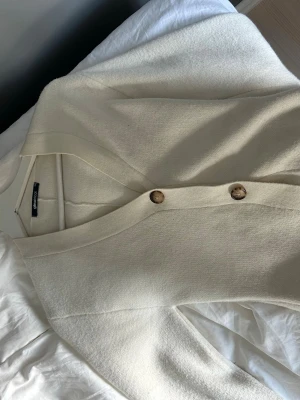 Vit kofta från Gina Tricot - Stilren vit kofta från Gina Tricot med v-ringning och stora bruna knappar framtill. Koftan har lång ärm och är tillverkad i ett mjukt, stickat material som känns skönt mot huden. Den formar sig efter kroppen och ger en elegant stil.  Perfekt att slänga över en topp för en enkel och snygg look. Använd 2 gånger