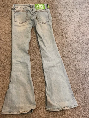 Ljusblå bootcut jeans med patch - Säljer ett par ljusblå low waisted bootcut jeans med utsvängda ben och klassisk femficksdesign. Jeansen har en grön patch med tryck bak i linningen och är tillverkade i denim. Perfekta för dig som gillar 2000 tals jeans 