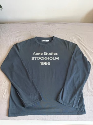 Mörkgrå långärmad Acne Studios tröja - Snygg mörkgrå långärmad tröja från Acne Studios med vit text 'Acne Studios STOCKHOLM 1996' på bröstet. Tröjan har rund halsringning och är gjord i mjuk bomull. Perfekt för en avslappnad och trendig look.