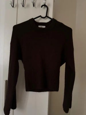 Mörkbrun stickad tröja Gina Tricot - Mysig mörkbrun stickad tröja från Gina Tricot i ullblandning. Tröjan har rund halsringning, långa ärmar och ribbstickade detaljer vid midjan och halsen. Perfekt för kalla dagar och enkel att matcha med jeans eller kjol.