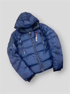 Polo Ralph Lauren RLX "Down Jacket" Jacka - Polo Ralph Lauren RLX Down Jacket | Modell: RLX Water-Resistant Down Jacket - Dunjacka med luva | Skick: Mycket fint skick / Sparsamt använd | Färg: Marinblå (Navy) med vita och röda detaljer | Material: Yttertyg 100% Nylon, Fyllning 75% Dun / 25% Fjäder | Nypris: ca 4 500 kr | Storlek: M - Passar även S | Hör av dig vid minsta fundering eller fråga 📦