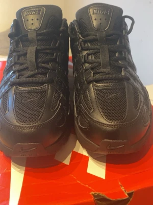 Svarta Nike sneakers i mesh och läder - Snygga helsvarta Nike sneakers med en mix av mesh och läderdetaljer. Skorna har klassisk snörning, diskreta swoosh-loggor och en chunky sula för extra streetkänsla. Perfekta för dig som gillar en clean och sportig look.