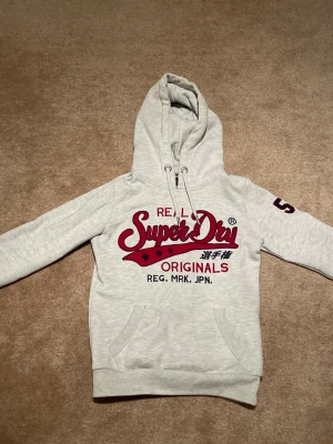 Superdry hoodie  - • ljusgrå Hoodie med röd text från superdry.          • Storlek XS.                                                                   •Nytt skick/mycket bra skick.                                    •skriv vid frågor, pris kan diskuteras👌🤝      