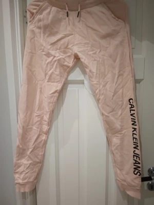 Beiga / rosa mjukisbyxor Calvin Klein Jeans, Strlk liten M  - Snygga beiga/rosa mjukisbyxor från Calvin Klein Jeans med svart logga längs ena benet. Byxorna har resår i midjan med dragsko, fickor på sidorna och muddar vid bensluten. Perfekta för chill dagar eller när du vill ha en avslappnad stil. Strlk Medium men är små i storleken och passar bäst någon med XS-S. Lite skrynkliga på bilderna men blir fina igen efter enkel strykning. Gott skick.