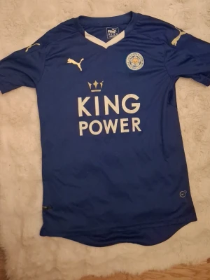 Blå Leicester City fotbollströja Puma - Säljer en blå Leicester City fotbollströja från Puma med klubbmärke på bröstet och King Power-tryck framtill. Tröjan har vita detaljer vid halsen och gula Puma-loggor på bröst och ärmar. Perfekt för fotbollsträning eller match.