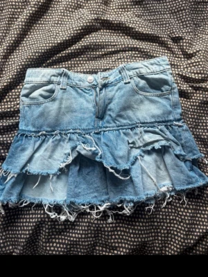 Blå volangkjol i jeans från Gina Tricot - Säljer en blå jeanskjol från Gina Tricot med volanger och råa kanter. Kjolen har klassisk femficksmodell, knapp och dragkedja framtill samt en cool, sliten look. Perfekt för dig som gillar en trendig och avslappnad stil.