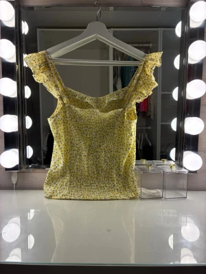 Gul blommigt linne  med volangdetaljer - Supersöt gul blus med små blommor och volangprydda axelband. Toppen har smockad överdel för skön passform och ärmlös design som känns luftig och somrig. Perfekt att styla med jeans eller kjol för en fräsch look.