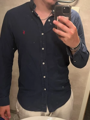 Mörkblå skjorta från ralph lauren - Mörkblå ralph lauren skjorta med röd logga, skjortan är i väldigt bra skick Brister: vissa knappar kan behövas sy fast mest men det är en easy fix. Passform: Slim fit, jag är 185 och den sitter perfekt