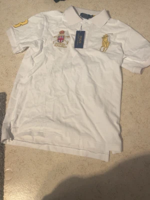 Vit pikétröja med brodyr Polo Ralph Lauren - Klassisk vit pikétröja från Polo Ralph Lauren med gul broderad logga på bröstet och ett broderat emblem. Tröjan har krage, knappar framtill och en gul detalj på ärmen. Perfekt till en stilren och sportig look.
