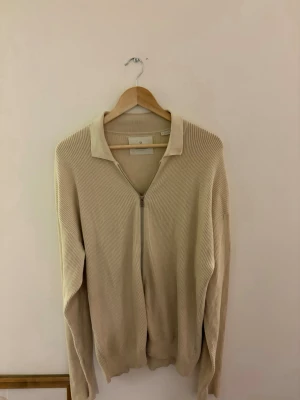 Beige ribbad ziptröja med krage - Stilren beige ziptröja med ribbad struktur och klassisk krage. Tröjan har långa ärmar och är tillverkad i ett mjukt stickat material som ger en avslappnad men snygg look. Perfekt att slänga på sig över en t-shirt för en enkel men trendig outfit.
