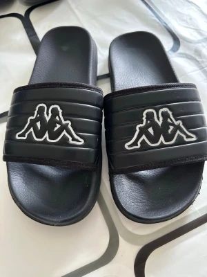 Svarta slides från Kappa - Säljer ett par svarta slides från Kappa med den klassiska loggan i vitt på ovandelen. Sandalerna har en bred rem med ribbad struktur och är tillverkade i syntetmaterial. Perfekta för sommaren eller att glida runt i hemma.